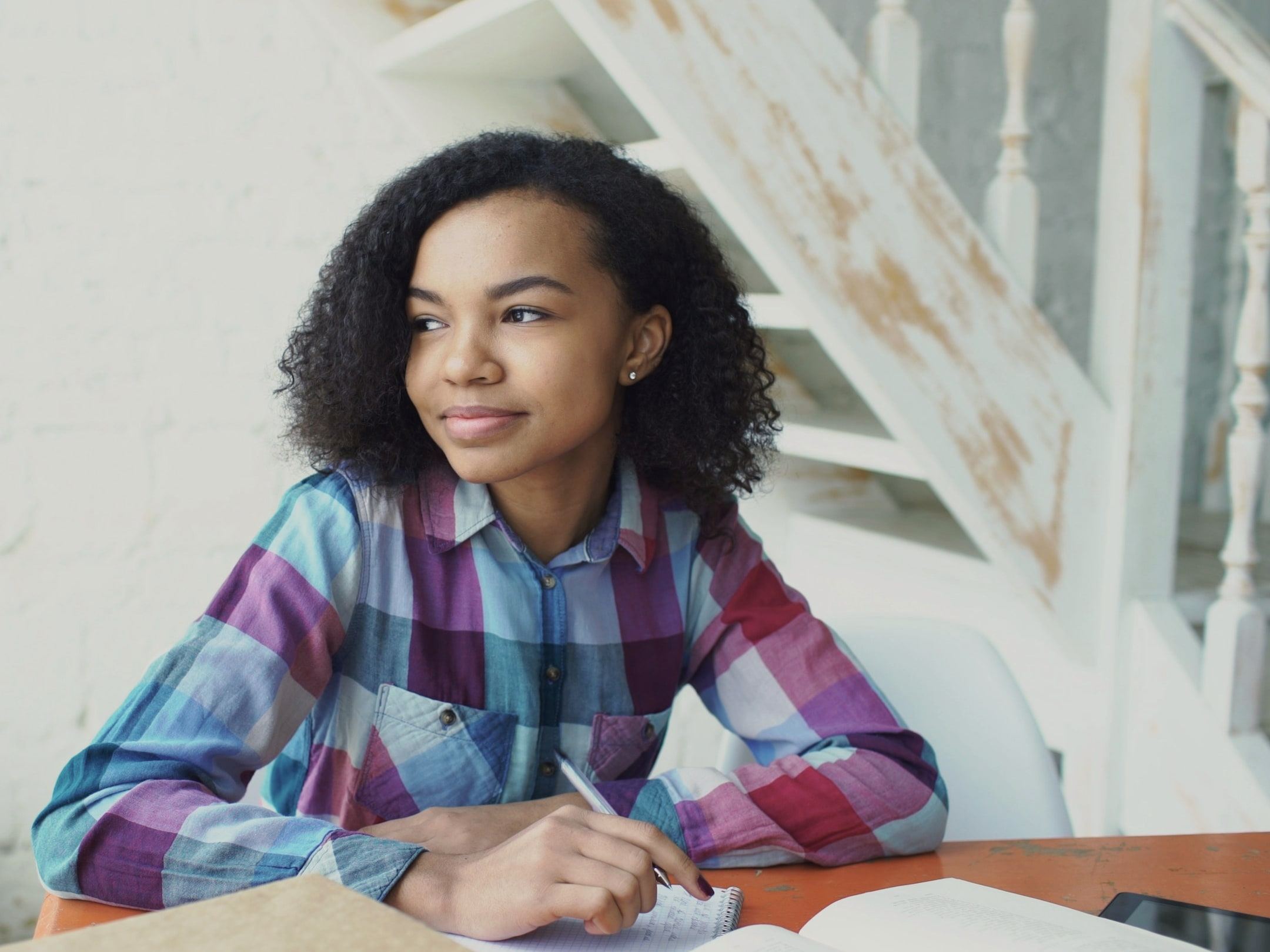A high school student pauses while writing notes at a sunlit table.